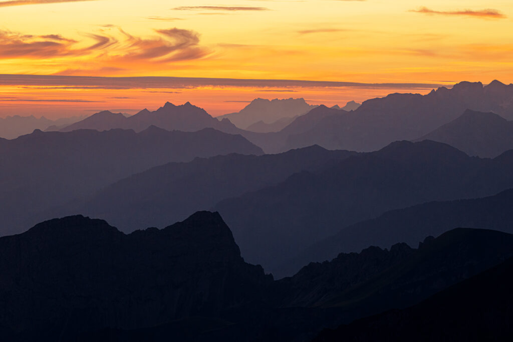 Mehrere Ebenen von Bergsilhouetten in Blau- und Violetttönen unter einem orange glühenden Abendhimmel