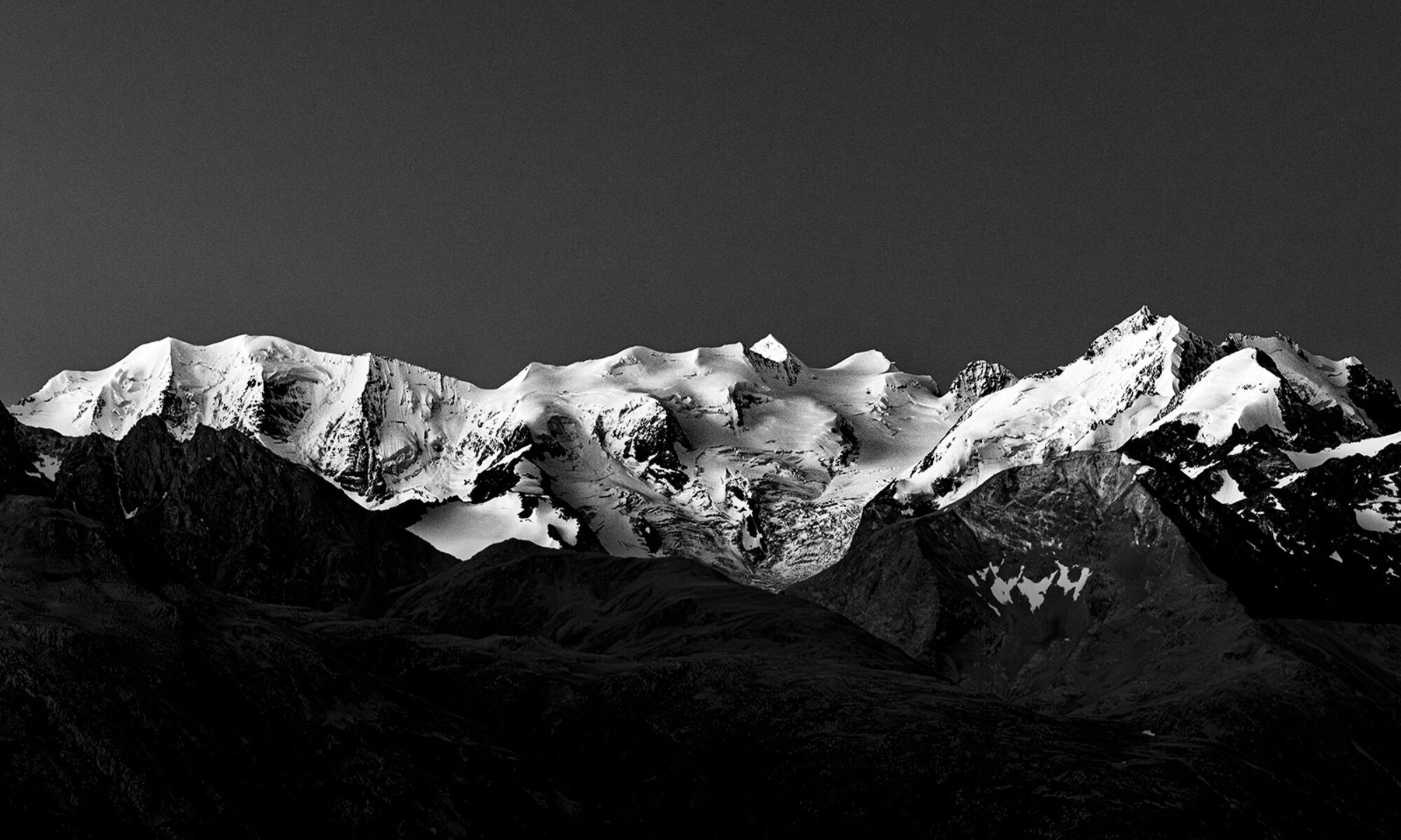 Panoramaaufnahme einer schneebedeckten Alpenkette in kontrastreichem Schwarz-Weiß vor einem dunklen Himmel.