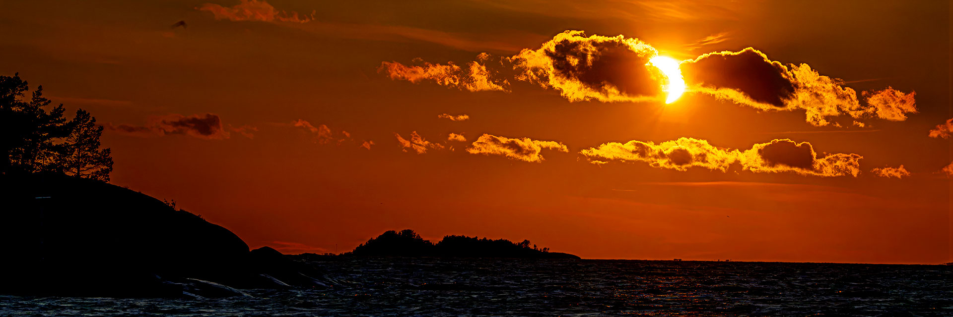 Panorama-Aufnahme einer partiellen Sonnenfinsternis im Abendlicht über dem Meer mit dunkler Küstensilhouette und Wolken.