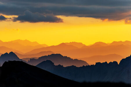 Farbenspiel im Hochgebirge: Spektakulärer Sonnenuntergang über den gestaffelten Gipfeln der Schweizer Alpen. Gestaffelte Bergsilhouetten in den Schweizer Alpen bei einem intensiv gelb und orange leuchtenden Sonnenuntergang unter einer dunklen Wolkendecke.