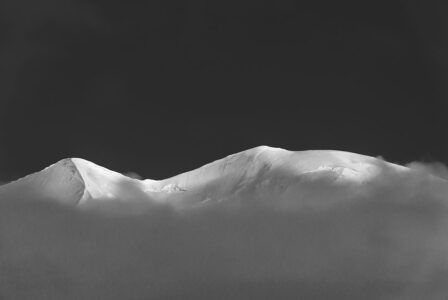 Über dem Nebelmeer: Die eisige Bergsilhouette des Piz Palü im Berninagebiet. Der schneebedeckte Gipfel des Piz Palü in den Schweizer Alpen in Schwarz-Weiss, der majestätisch aus einem dichten Wolkenmeer hervorragt.